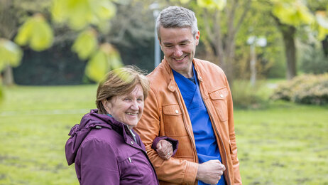 Ein Klinikmitarbeiter geht mit einer Patientin im Garten spazieren